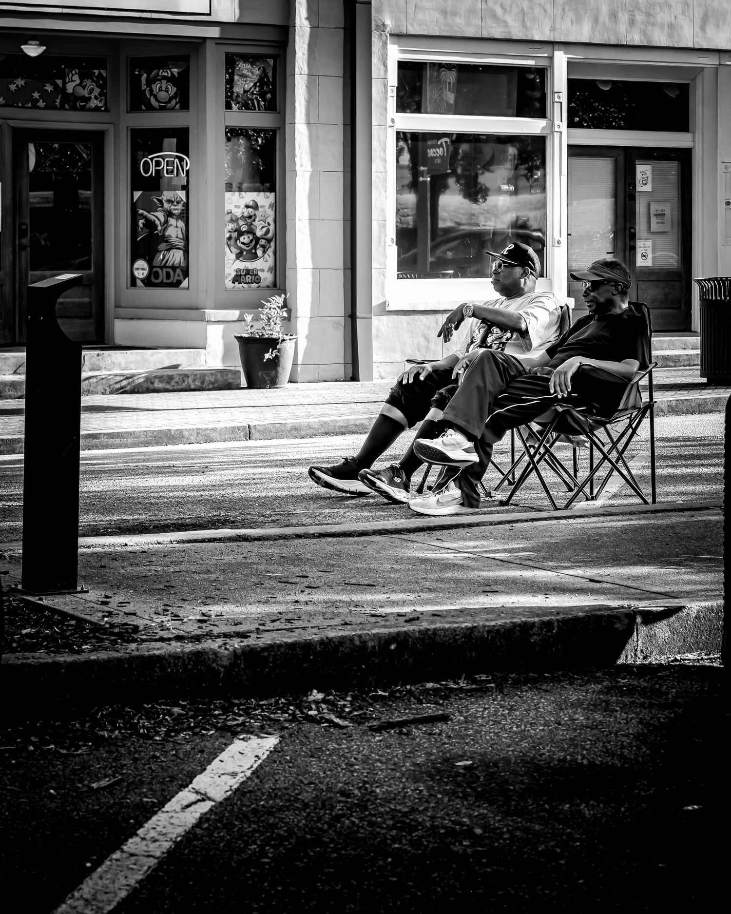 Two people sitting in folding chairs on a sidewalk outside a storefront in a black and white street photo.