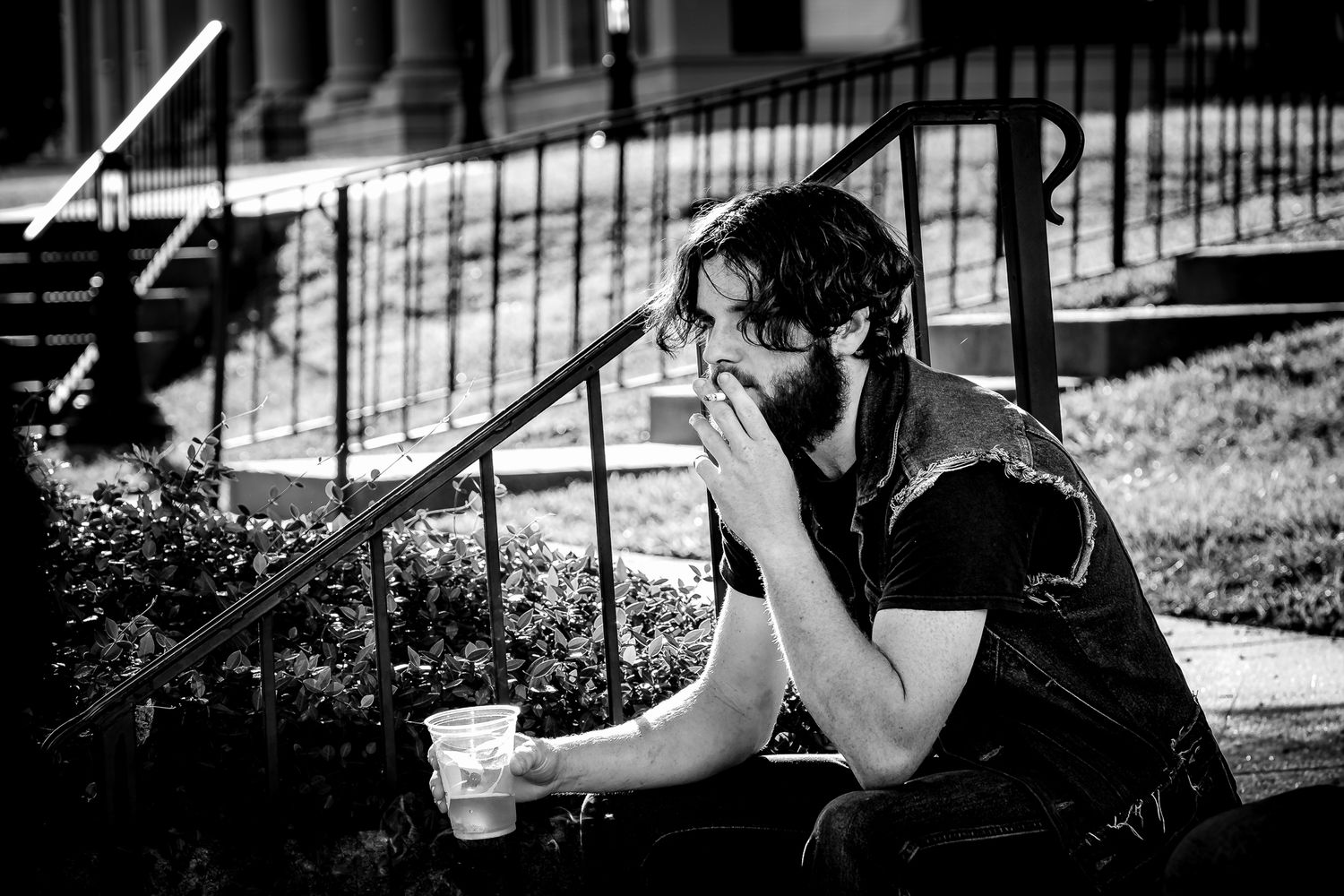 Black and white photo of a bearded man sitting on city steps, smoking and holding a cup outdoors.