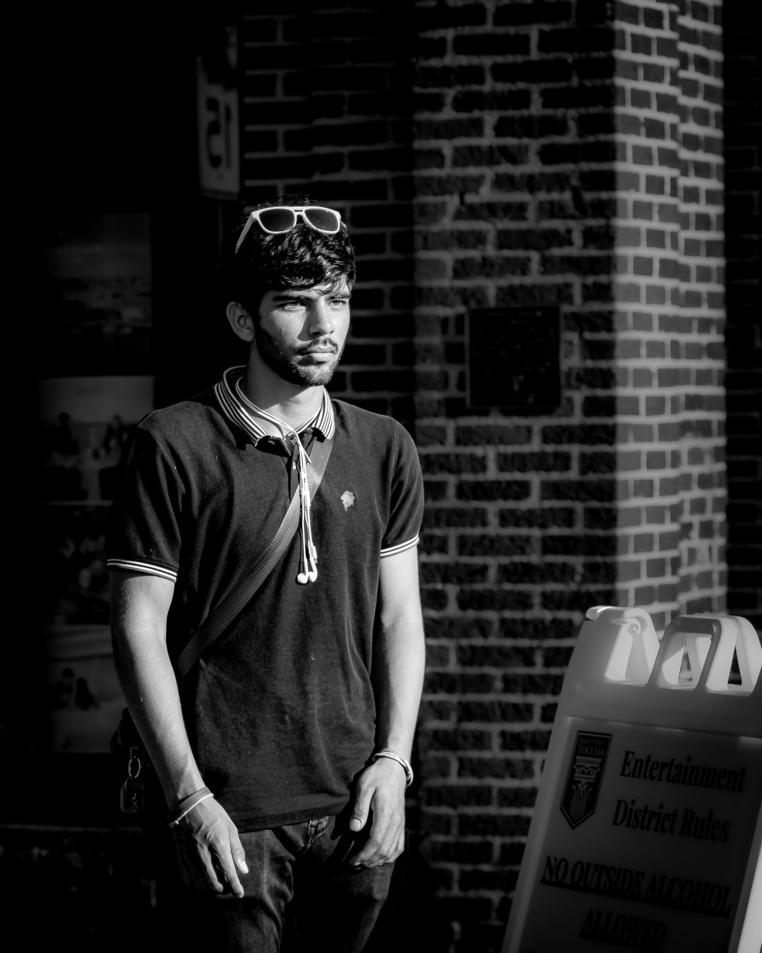 Black and white portrait of a young man with sunglasses on head, wearing a polo shirt, standing near a brick wall.