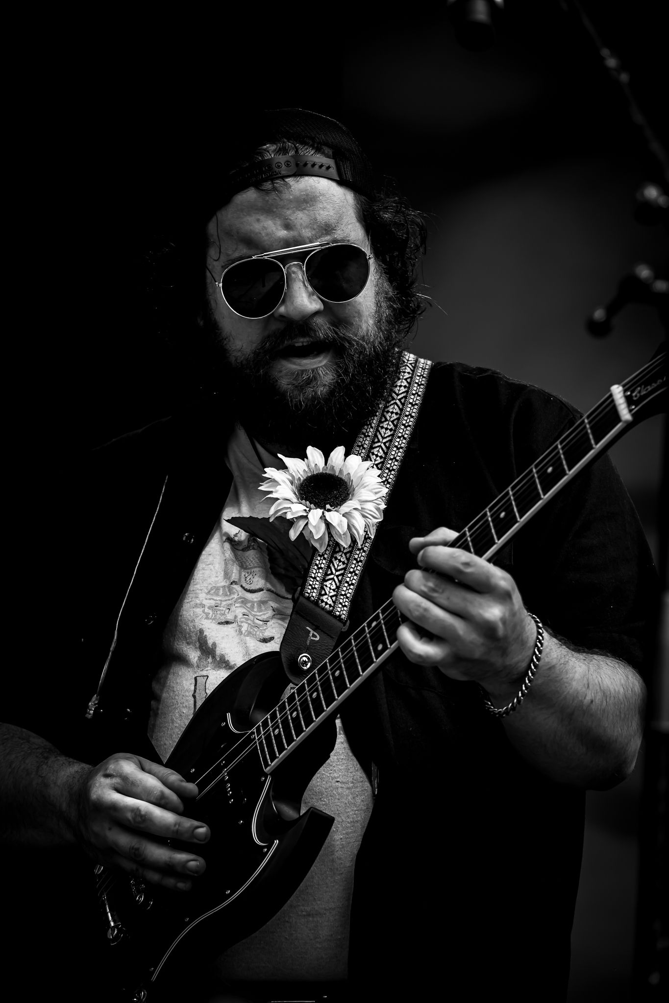 Black and white photo of a bearded male guitarist with sunglasses and a sunflower on his guitar strap.