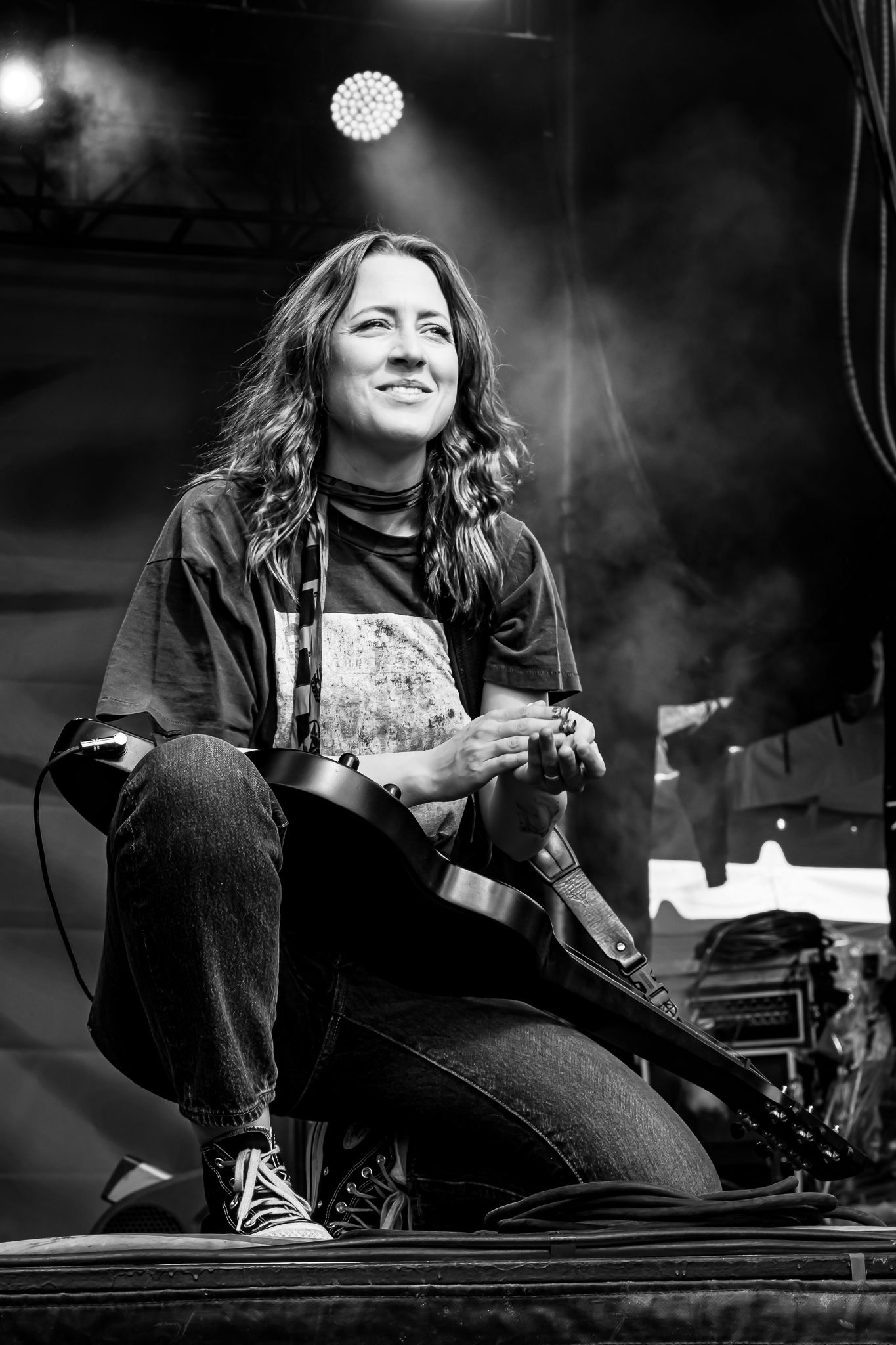 Female guitarist sitting on stage edge smiling, captured in black and white concert photo.