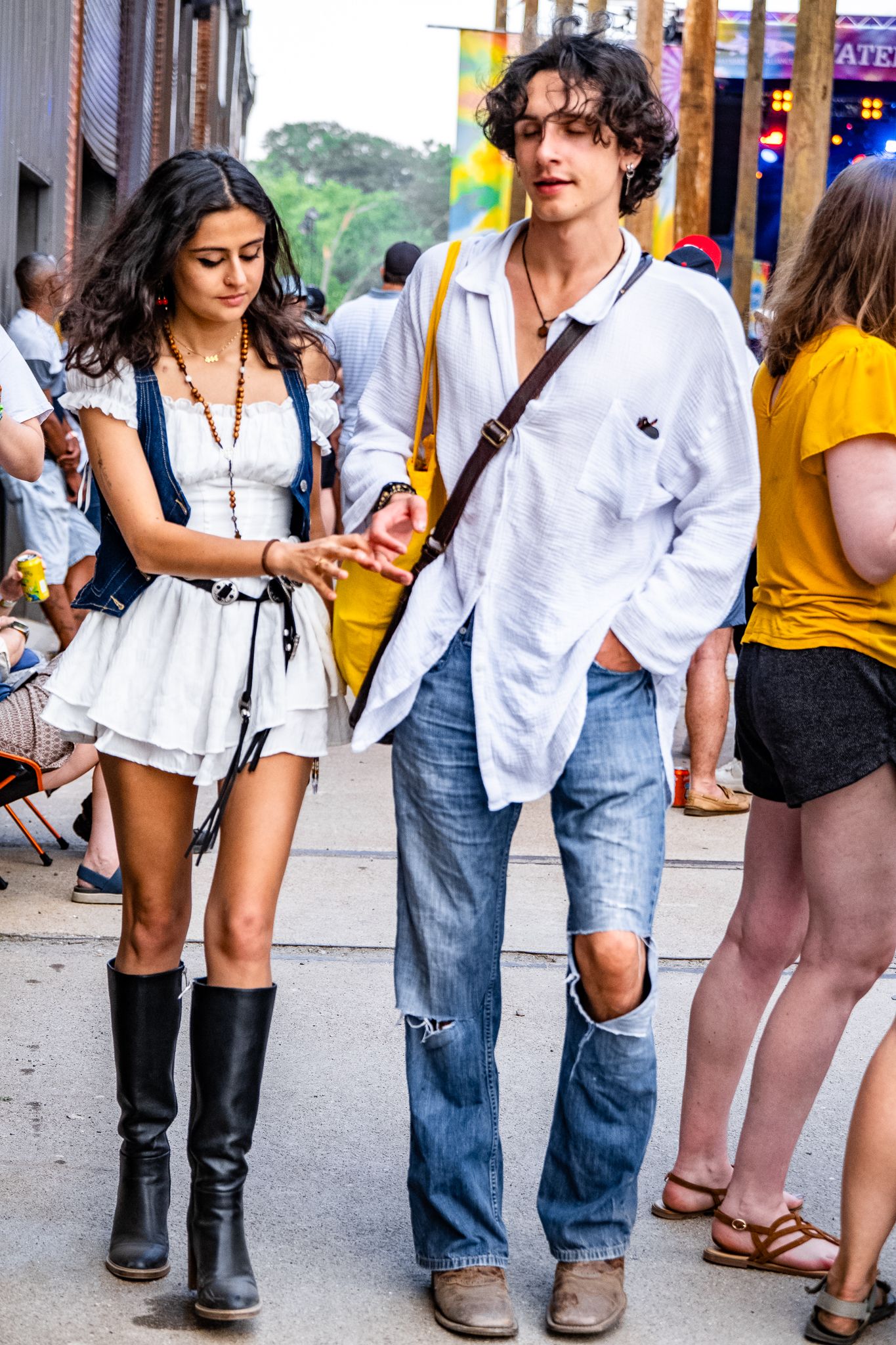 A young couple walks together at an outdoor festival, woman in white dress with black boots, man in white shirt.