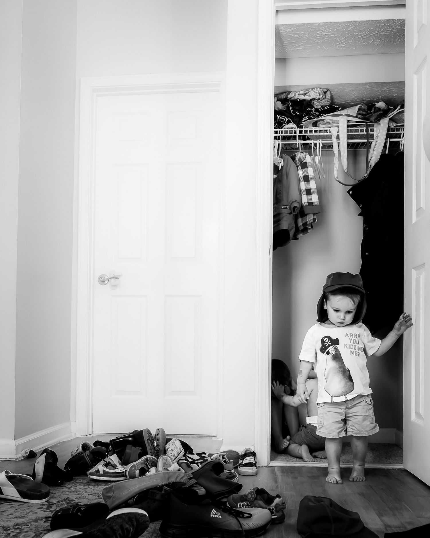 Black and white photo of a young child standing in an open closet doorway, with shoes scattered on the floor nearby.