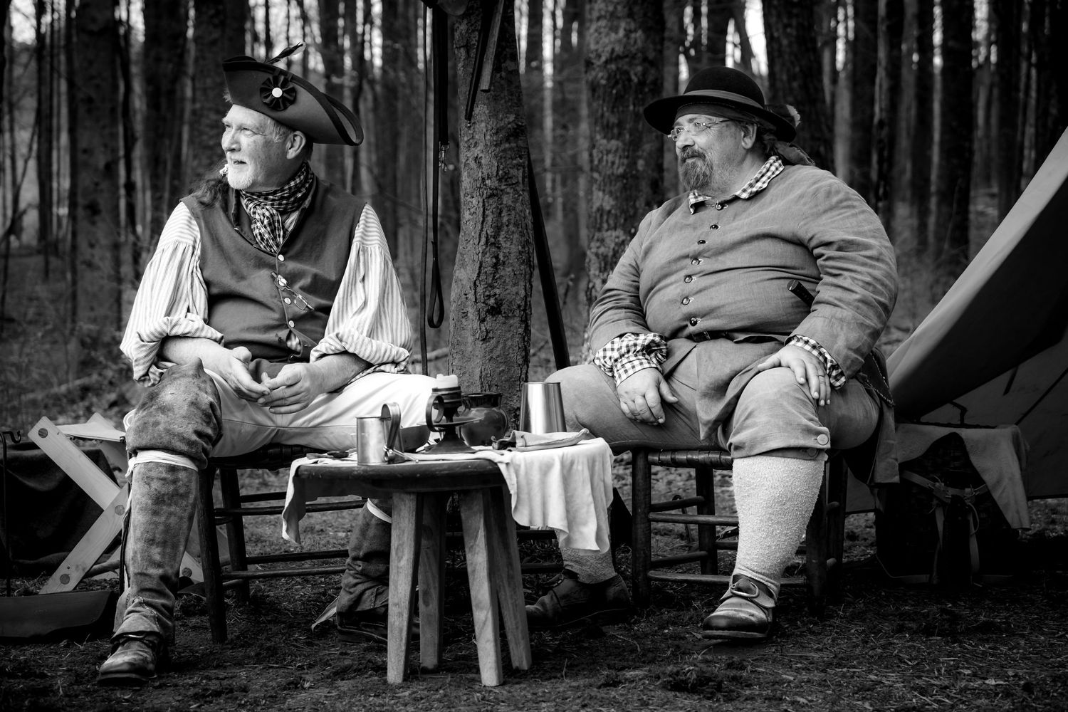 Two men in colonial frontier costumes rest at woodland camp, black and white historical reenactment.
