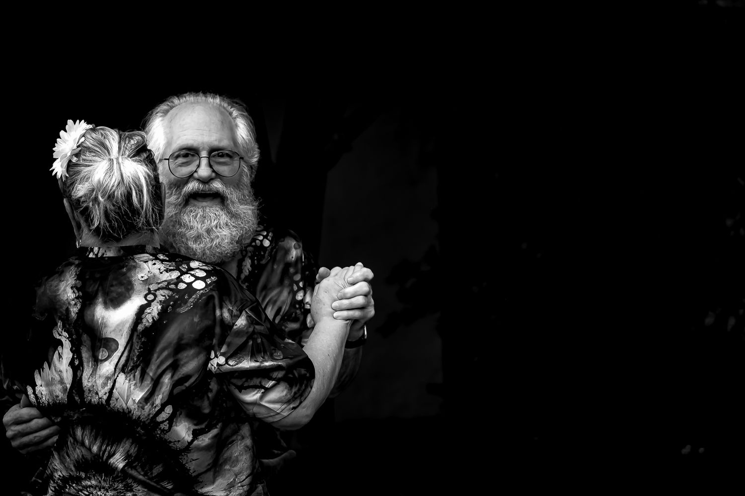 Black and white photo of a bearded man with glasses hugging a small dog, smiling warmly in dark background.