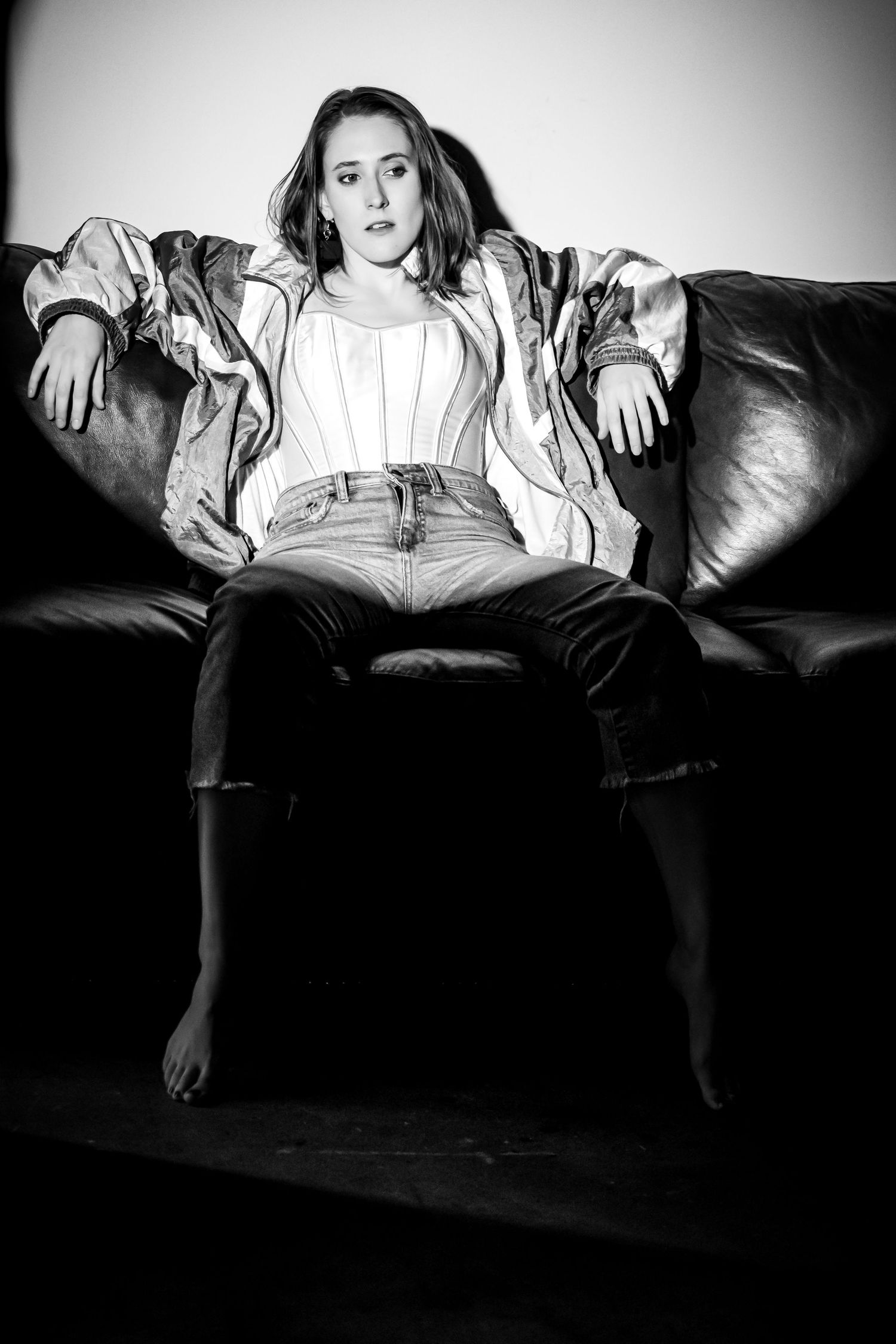 Young woman in jacket and jeans sits confidently on leather sofa in black and white portrait photo.