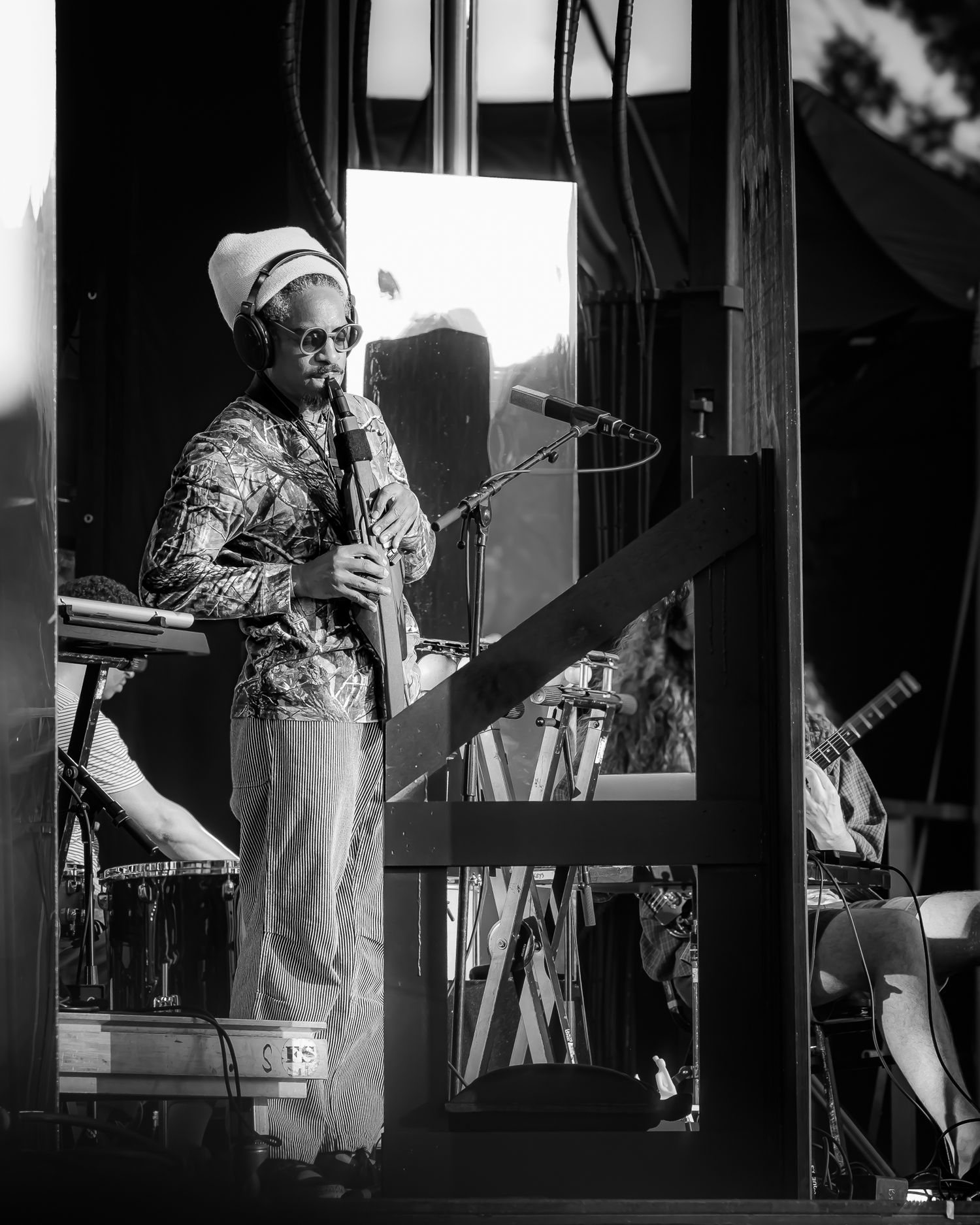 Black and white concert photo of a performer in headphones and white hat playing instrument on outdoor stage.