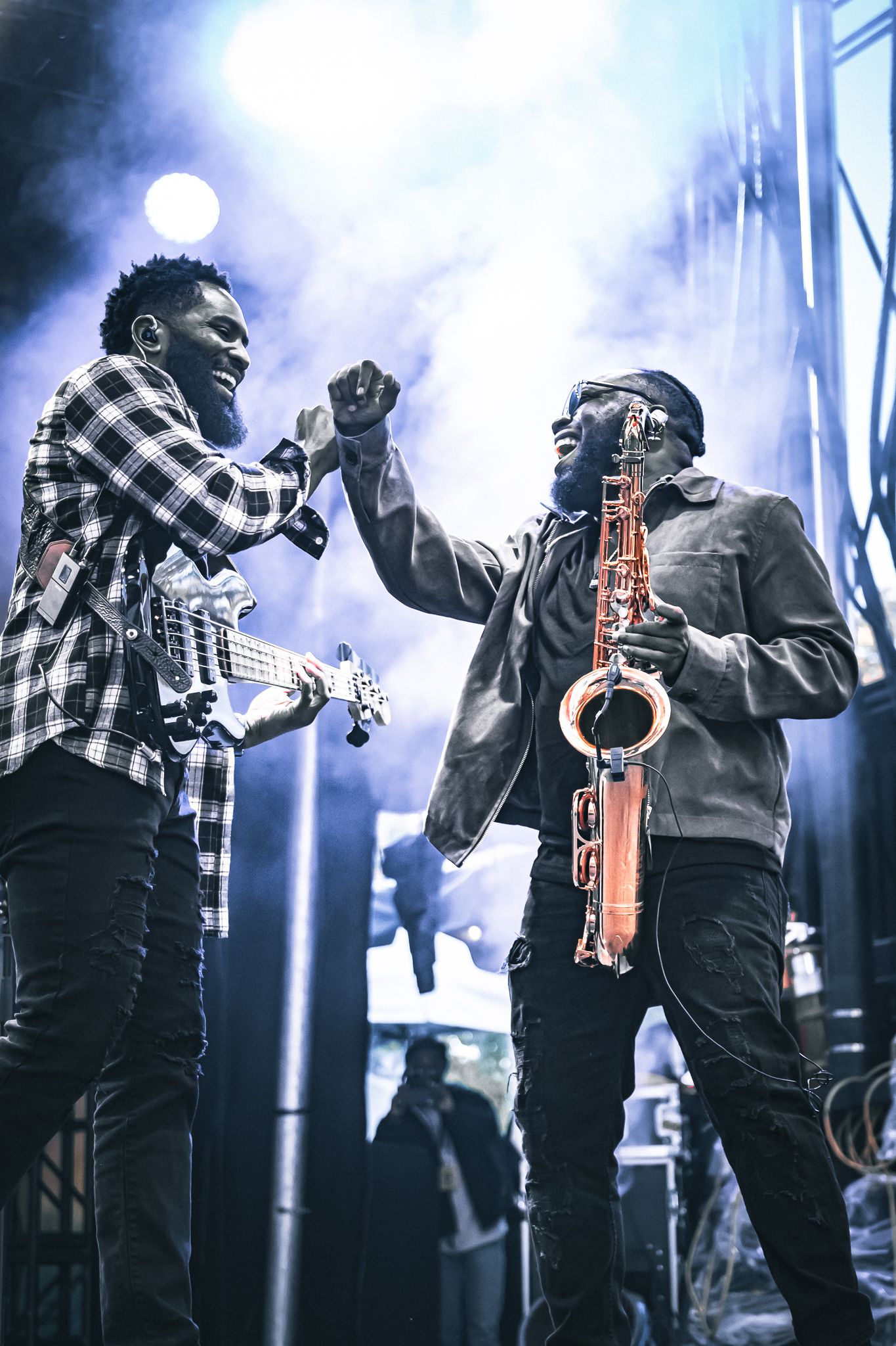 Two musicians celebrate on stage, one holding a saxophone, sharing a joyful fist bump during a live performance.