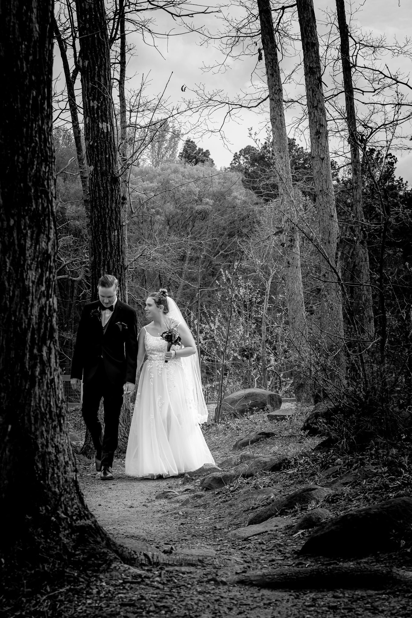 Elegant black and white wedding portrait of bride and groom strolling together along a scenic wooded forest trail.