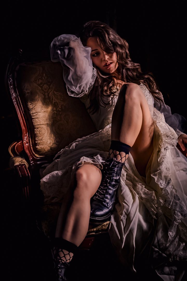 Young woman in a white bridal gown with lace-up boots poses dramatically in an ornate vintage armchair.
