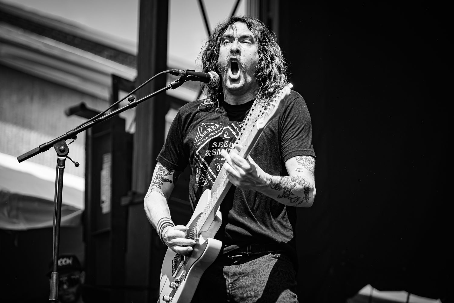 Tattooed male guitarist screaming into microphone while performing on stage in black and white concert photo.