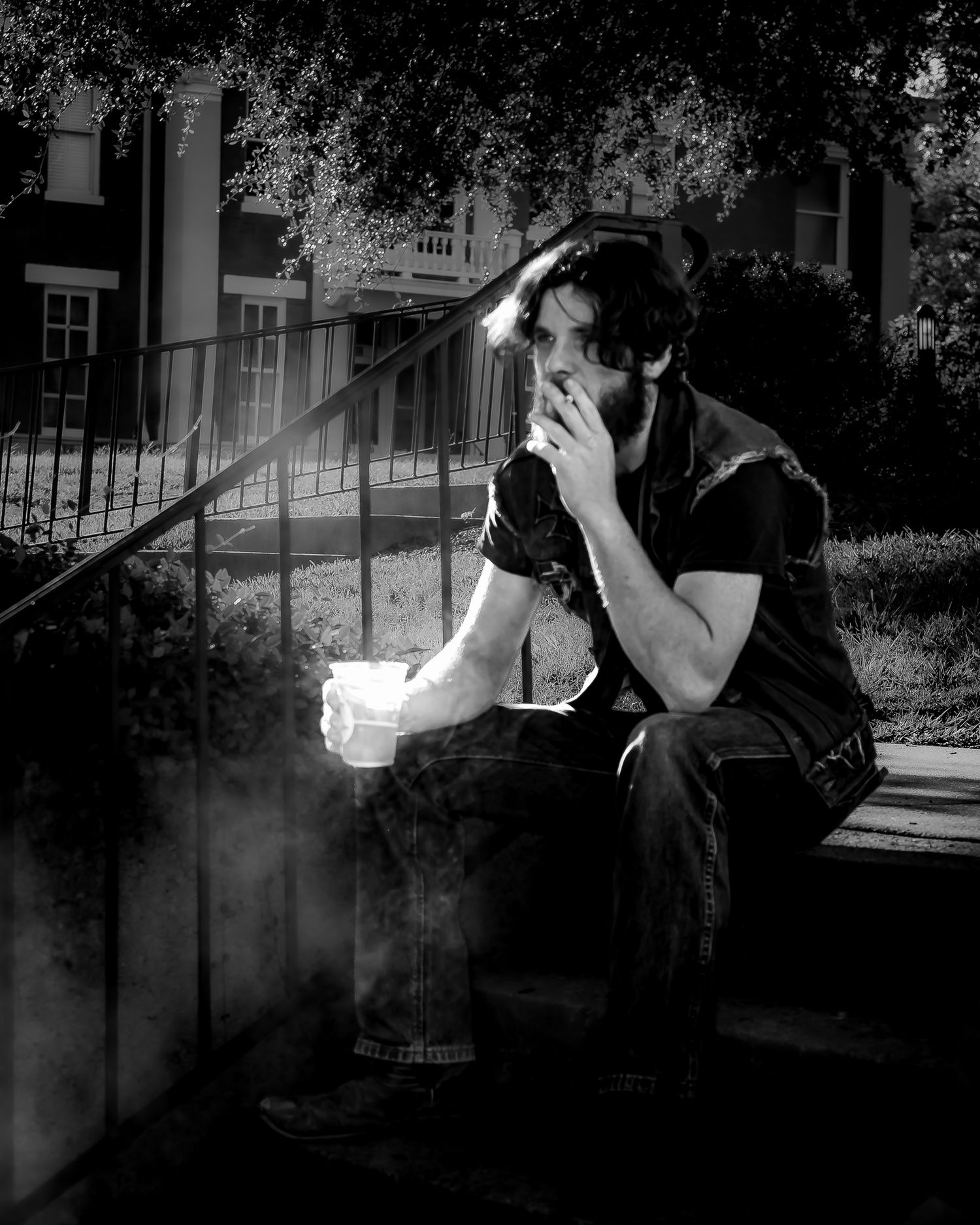 Black and white photo of a man sitting on steps outdoors, smoking and holding a cup.