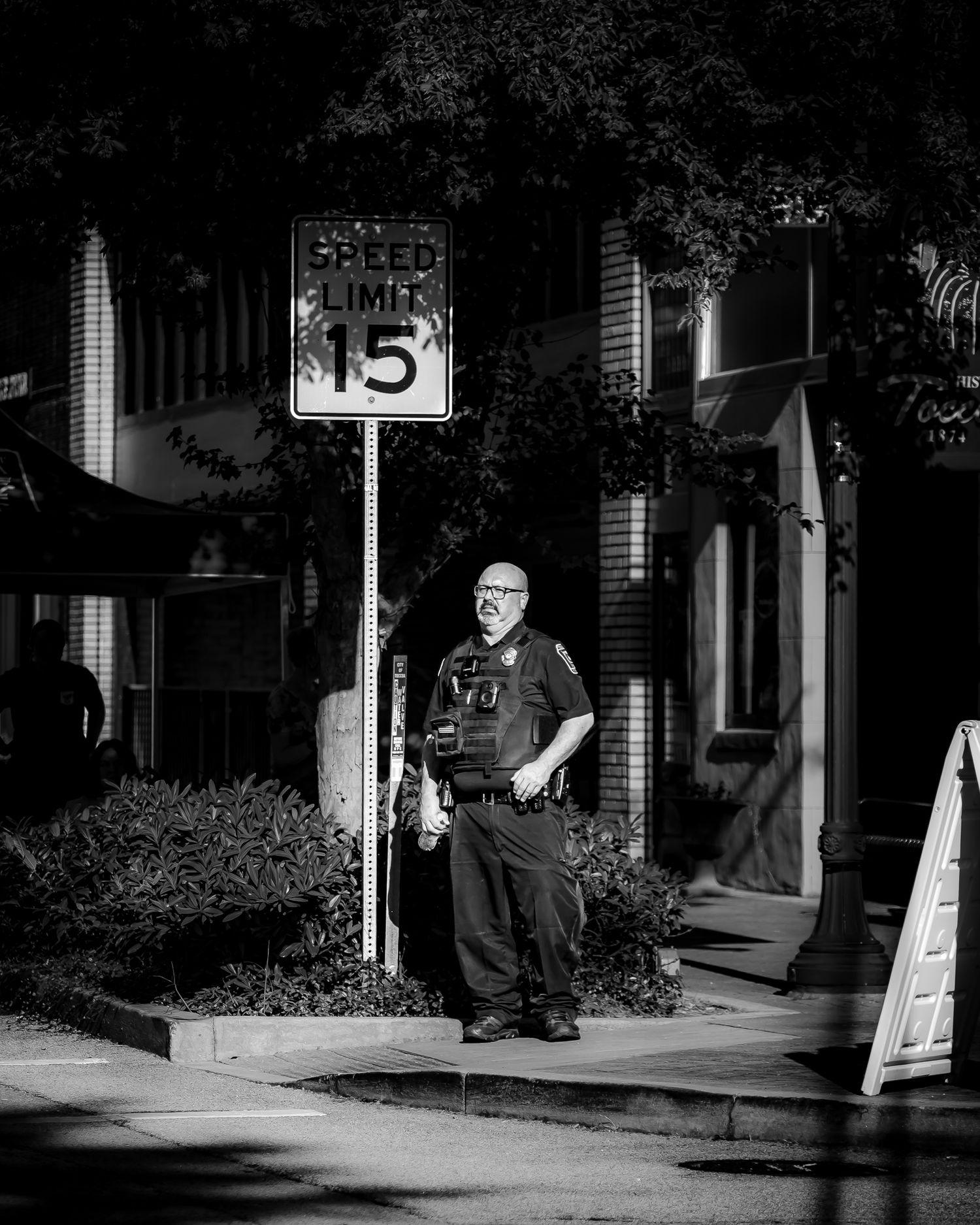 Black and white photo of a police officer standing beside a speed limit 15 sign on a sidewalk.