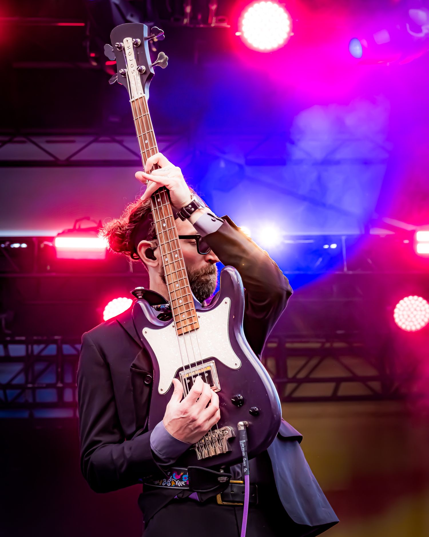 Bass guitarist raising instrument overhead under colorful stage lights at live music festival.