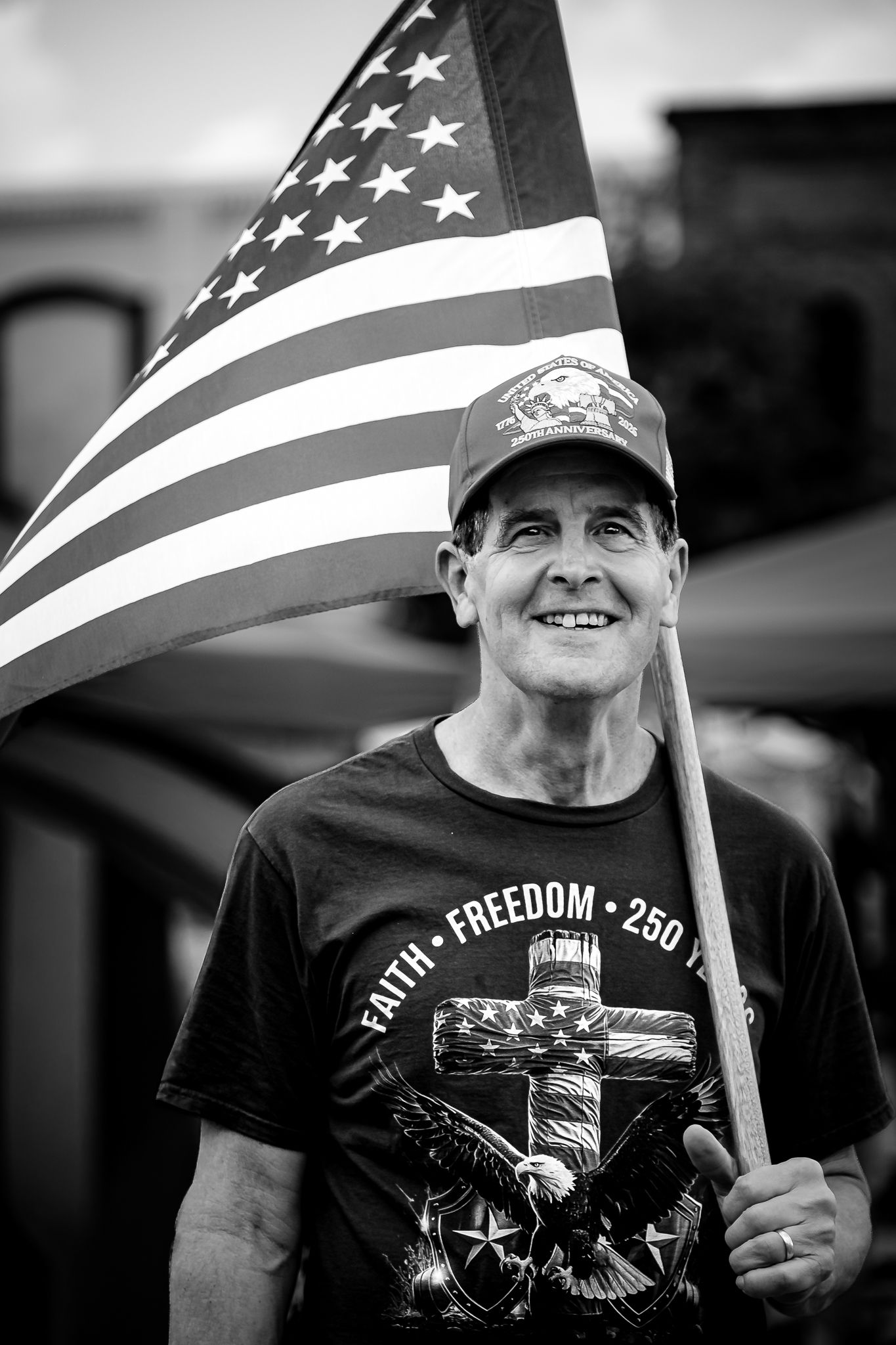 Black and white photo of smiling man holding American flag, wearing Faith Freedom t-shirt and cap at outdoor event.