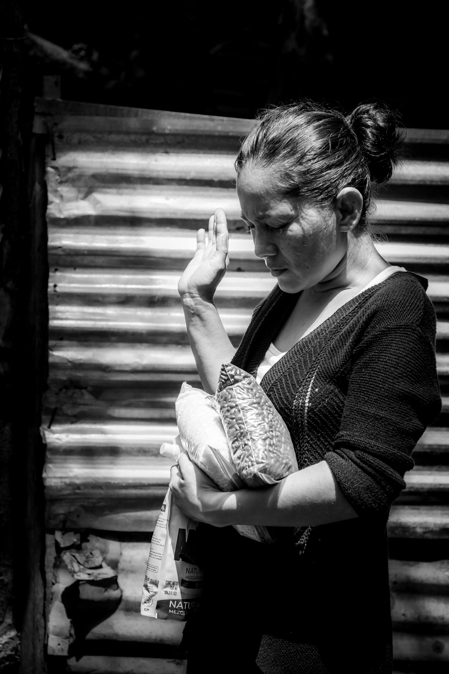 Black and white portrait of a woman holding a bundle, raising her hand near a corrugated metal wall.
