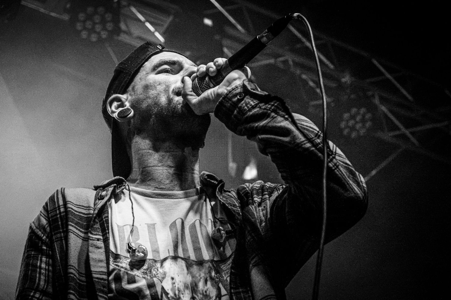 Black and white close-up of a male vocalist singing intensely into a microphone wearing a plaid shirt on stage.
