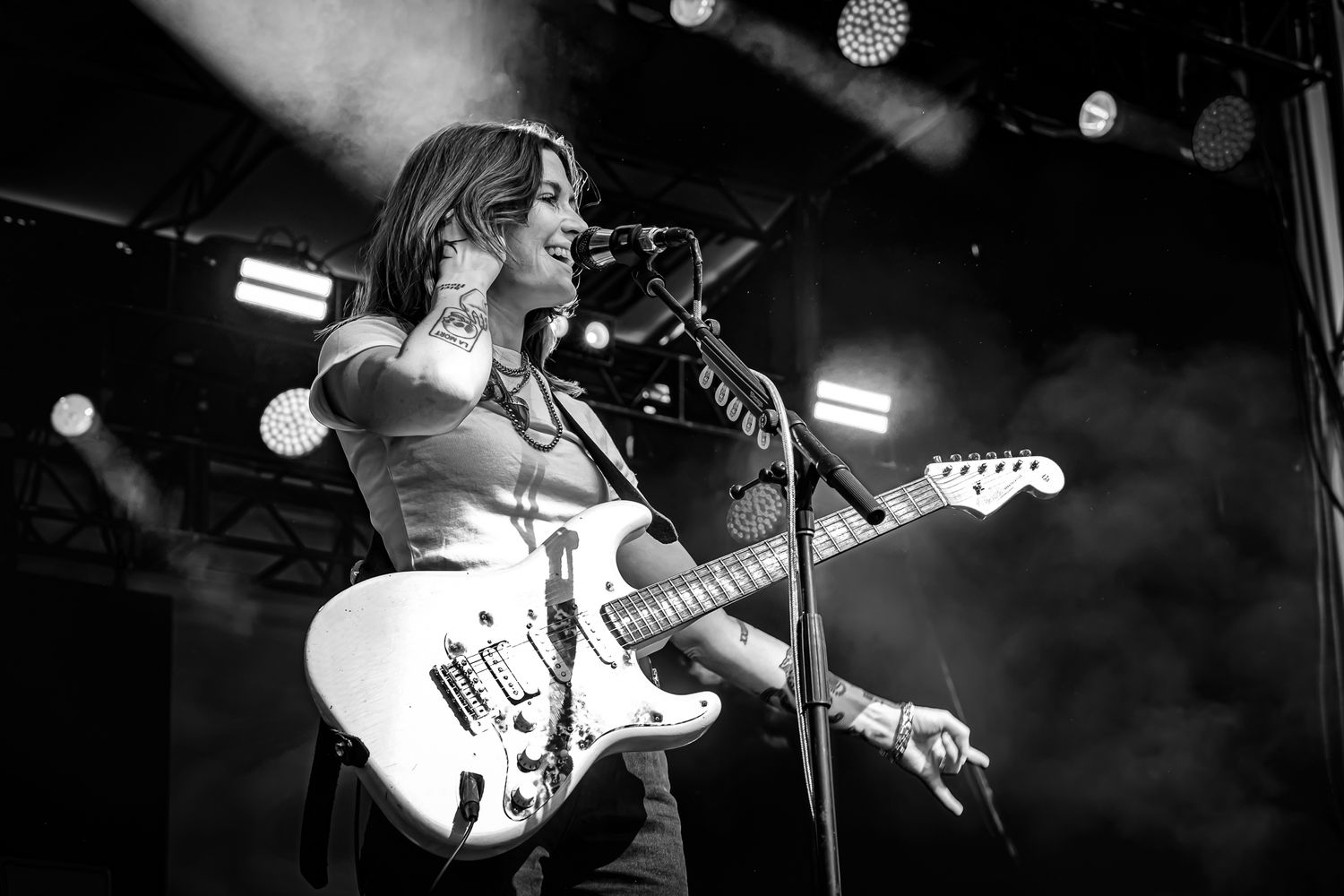 Tattooed female guitarist playing white electric guitar on smoky stage in black and white concert photo.