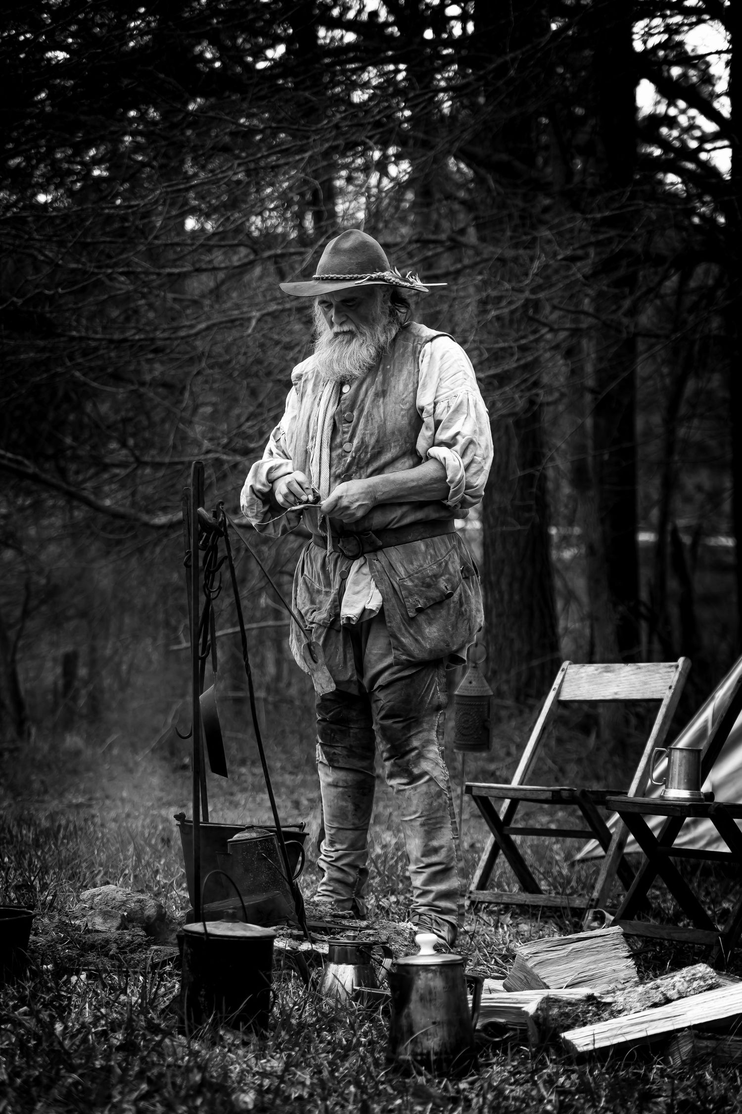 Bearded mountain man in frontier clothing tends campfire in forest, black and white historical reenactment.