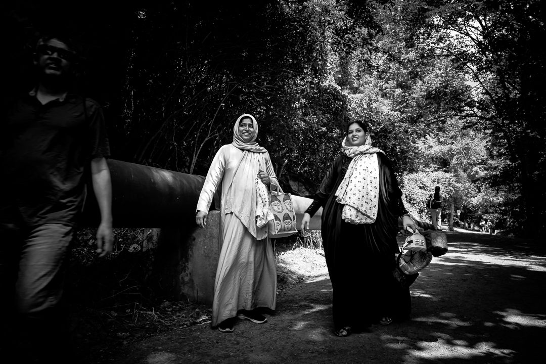 Two women in headscarves walking along a tree-lined path carrying bags.