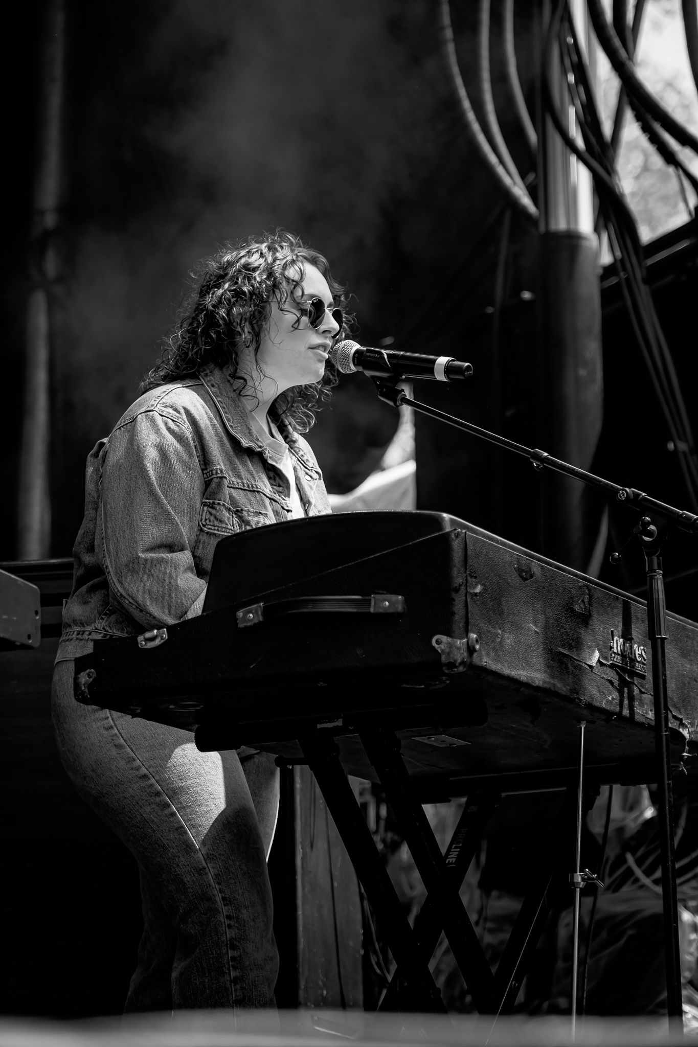 Curly-haired musician playing keyboard and singing into microphone on stage in black and white.