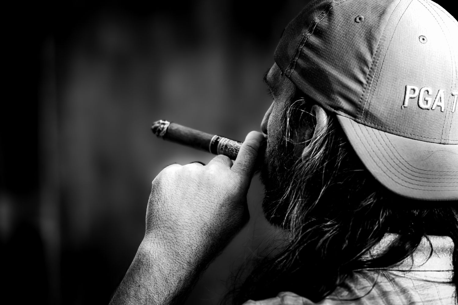 Black and white photo of a man wearing a PGA cap smoking a cigar, viewed from behind.