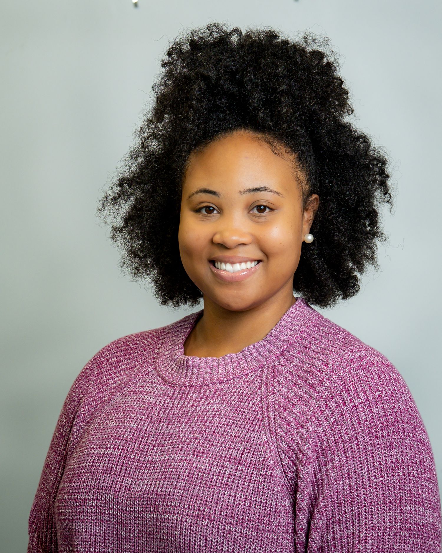 Smiling young woman with natural curly hair wearing a pink knit sweater against a gray background. Headshot