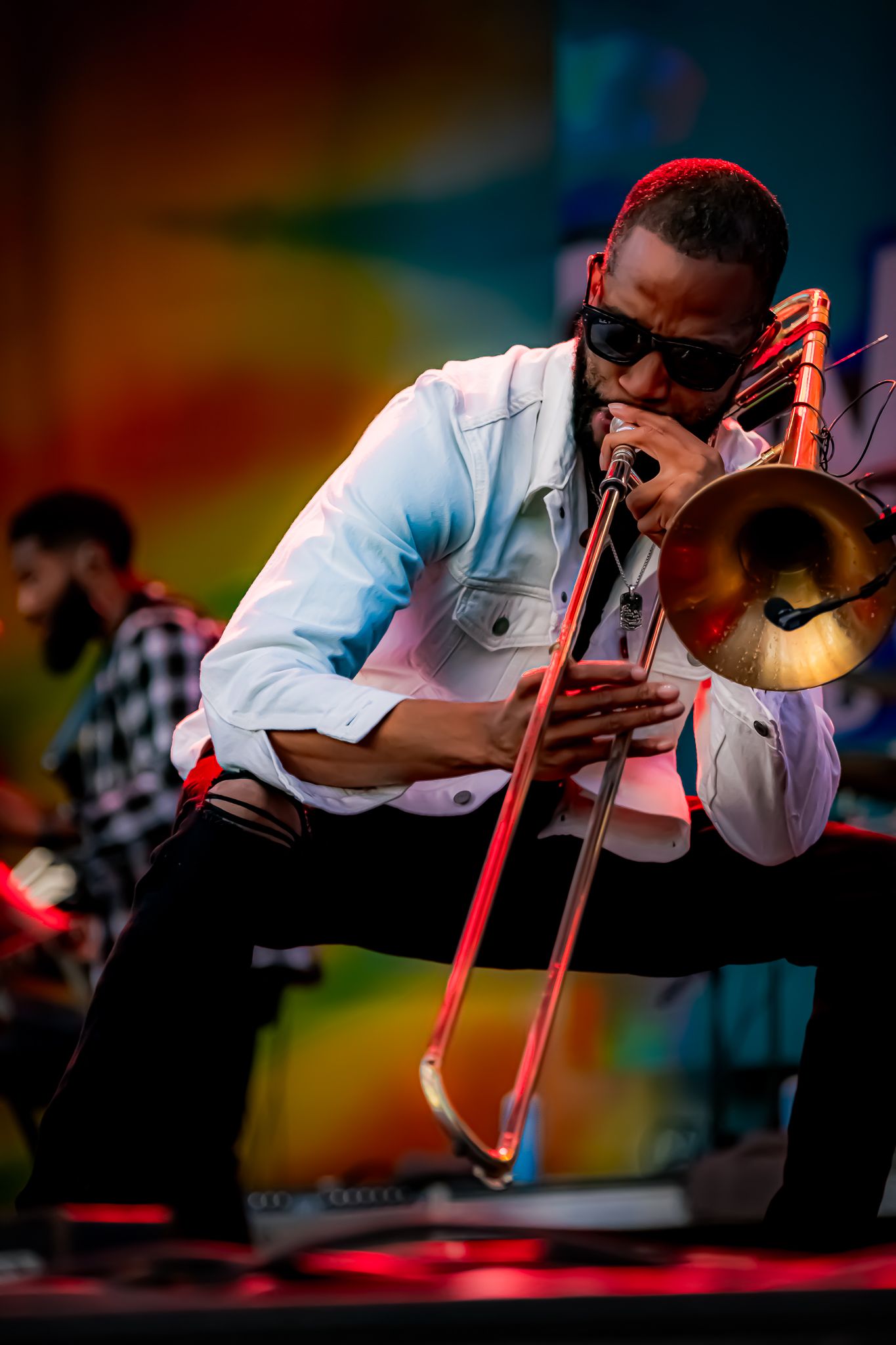 Trombone player performing on stage outdoors at a colorful music festival, wearing sunglasses and a white jacket.