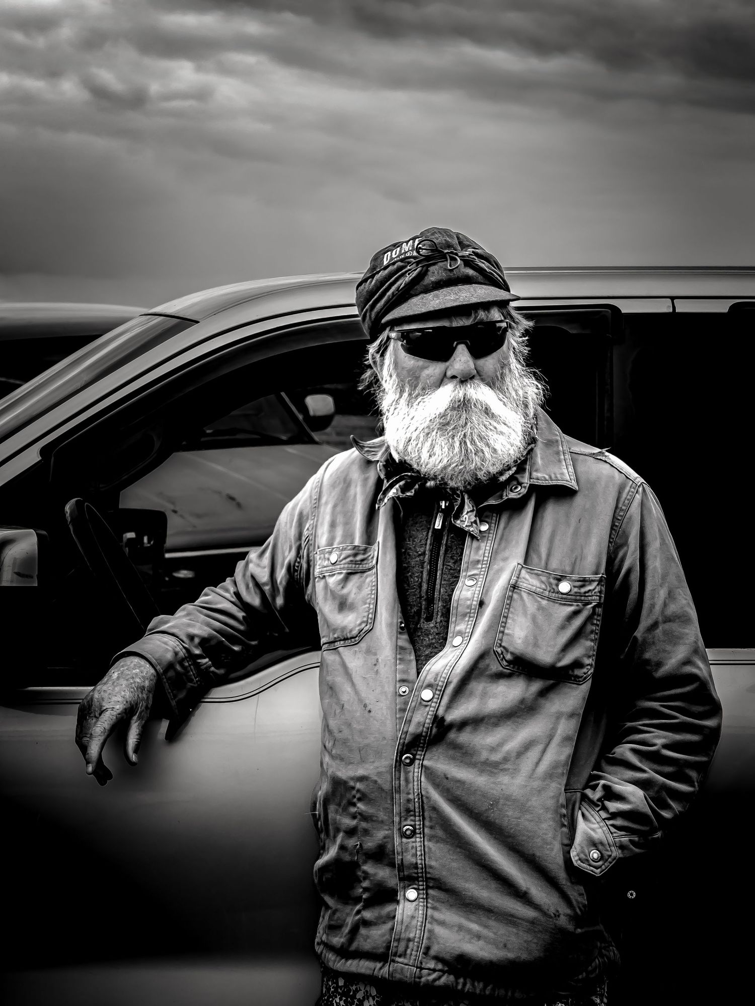 Rugged older man with white beard and sunglasses leaning on a truck under dramatic cloudy skies in black and white.