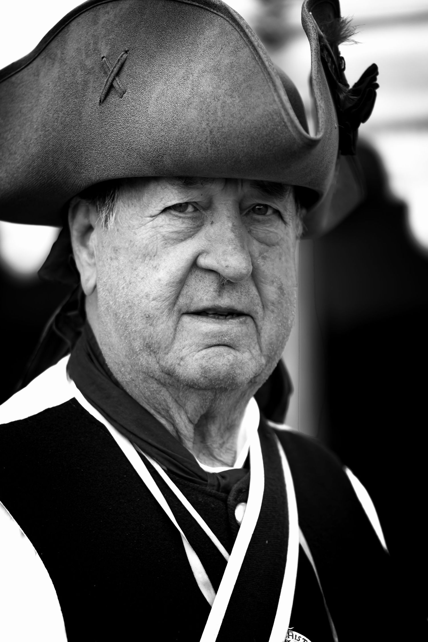 Black and white close-up portrait of an elderly man wearing a traditional tricorn hat and colonial-era costume.