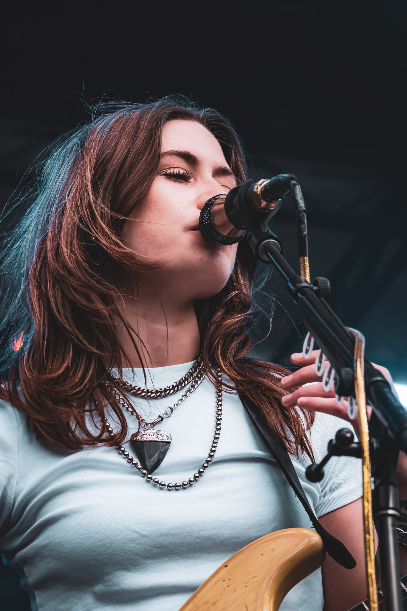 Female musician singing into microphone while playing guitar, wearing white tee and layered necklaces.