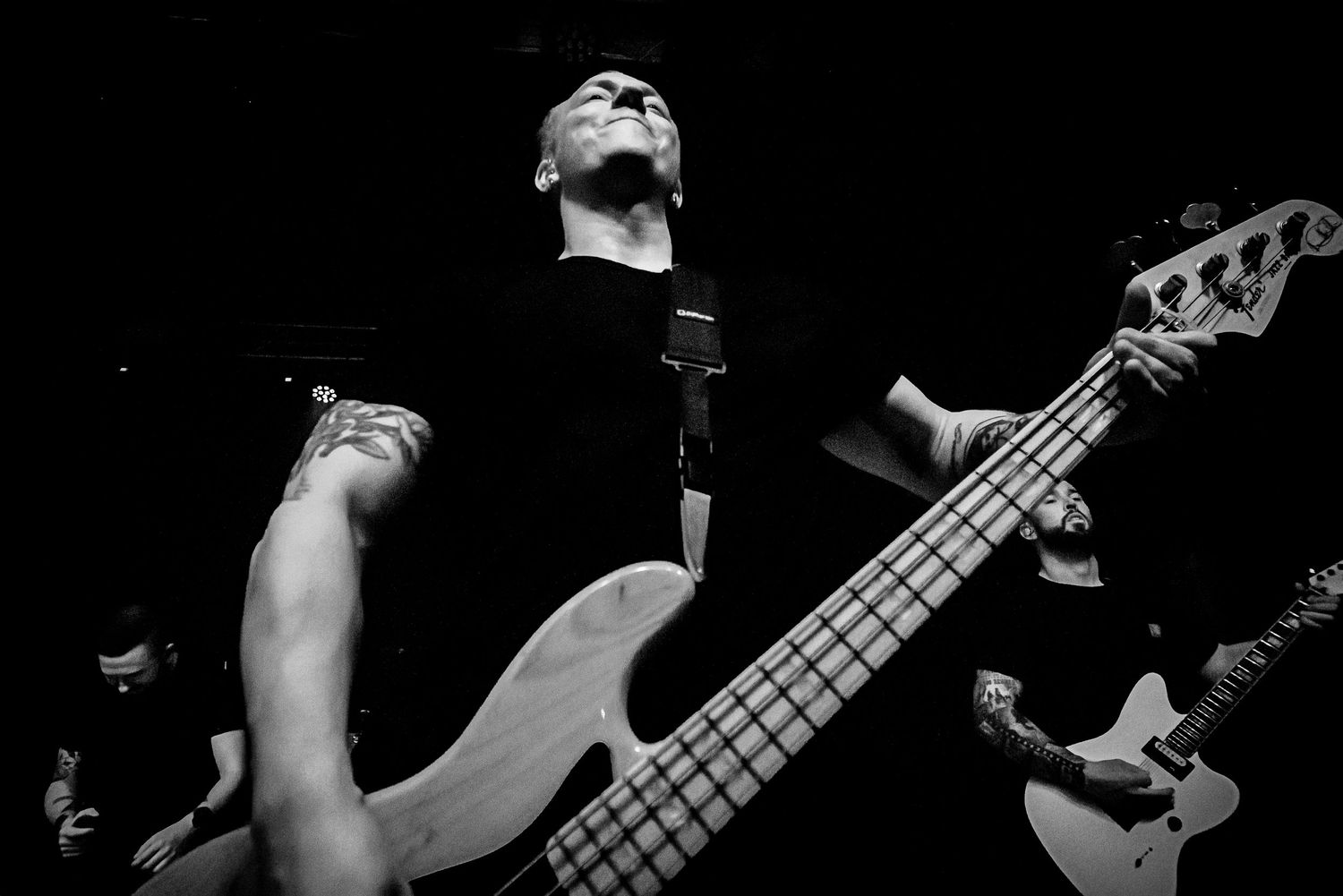 Black and white concert photo of a bassist playing a white five-string bass guitar on stage with band members.