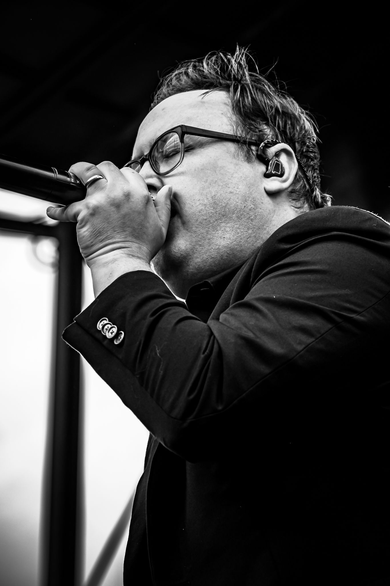 Black and white close-up of male vocalist singing passionately into microphone wearing glasses.