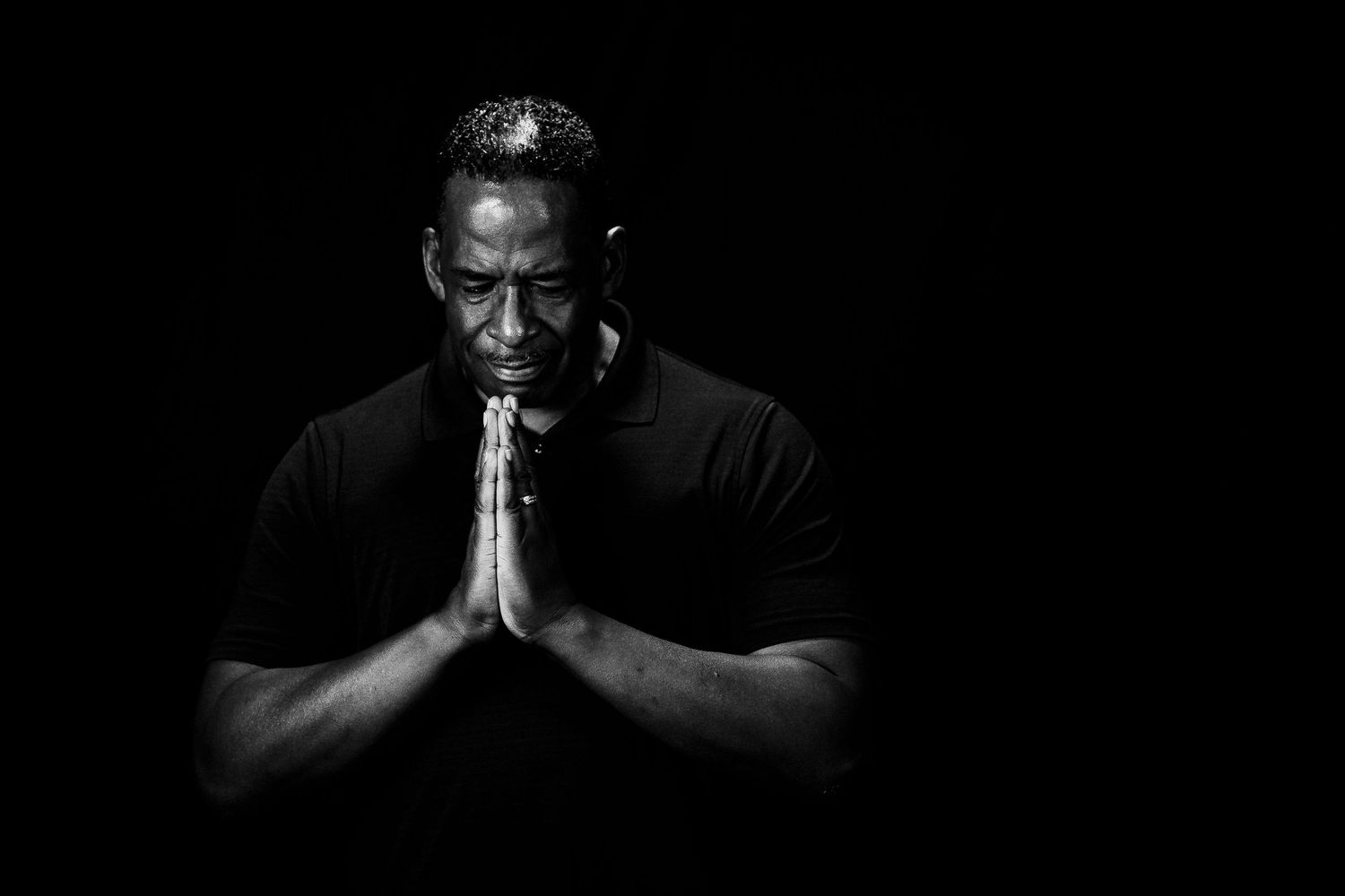 Black and white portrait of a man in prayer pose with hands clasped, dramatic low-key lighting against dark background.