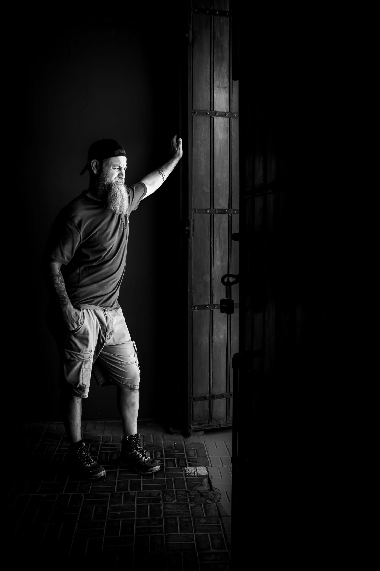 Bearded man in cap and shorts leaning against a rustic wooden door in dramatic black and white photography.