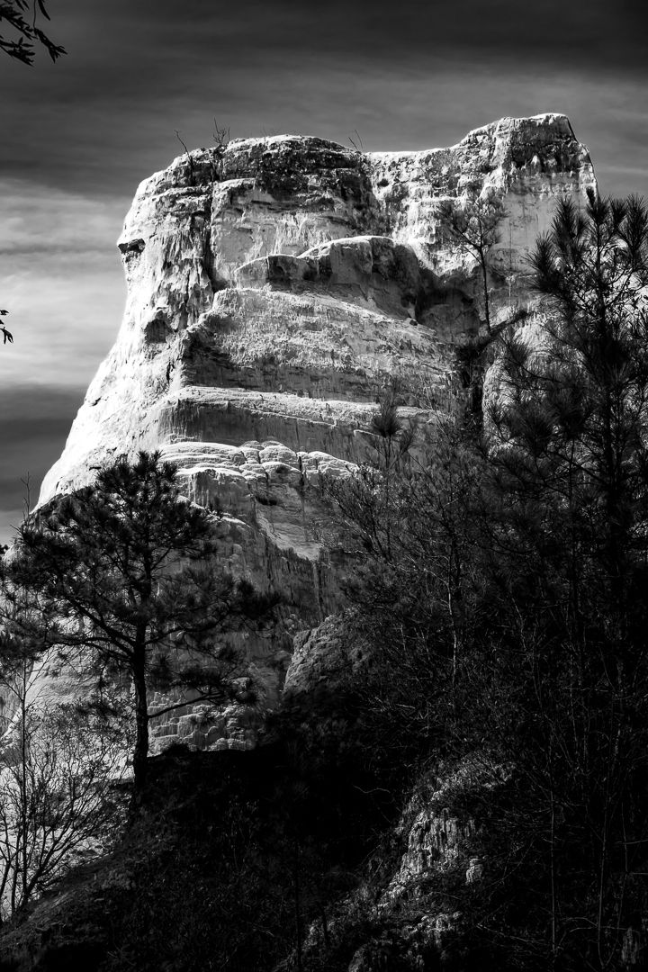 Black and white photo of a dramatic stepped sandstone rock formation towering above pine trees against a cloudy sky.