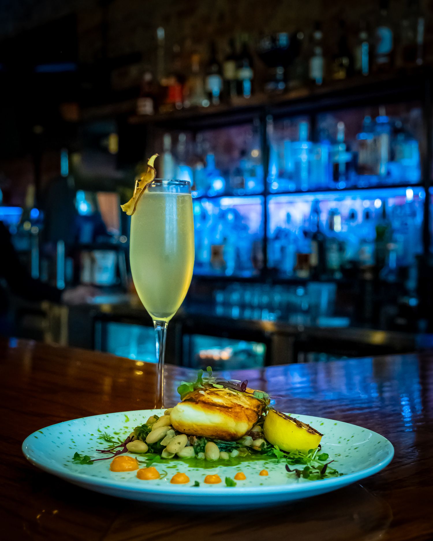 Elegant champagne flute cocktail beside a gourmet fish dish on a white plate, set against a glowing blue bar backdrop. Food. Restaurant.