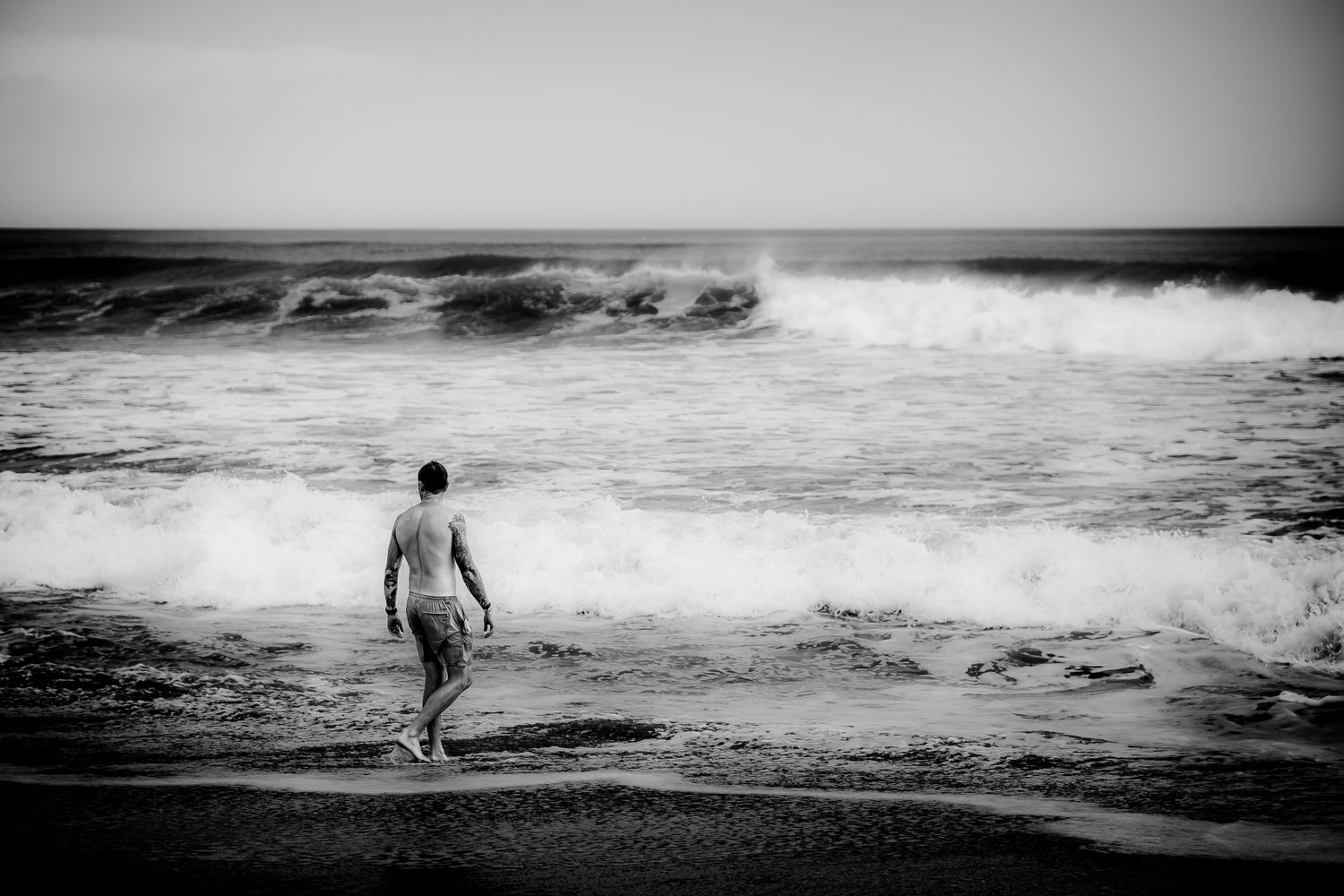 Lone man walks toward crashing ocean waves on dark beach in black and white photography. Landscape, travel documentary, travel