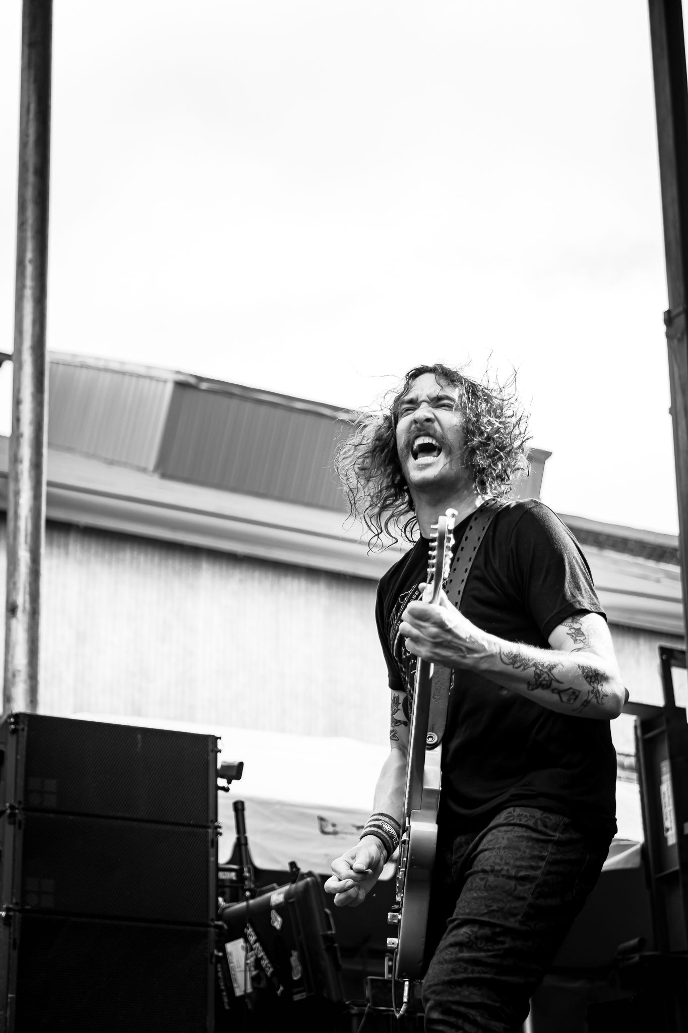 Long-haired tattooed guitarist screaming passionately while performing on outdoor stage in black and white.