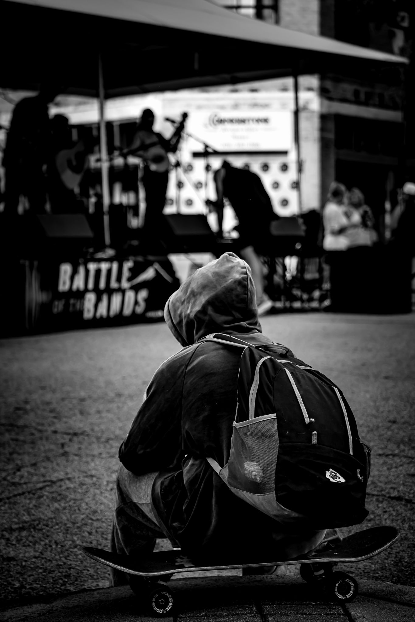 Person sitting on skateboard watching Battle of the Bands outdoor concert, black and white photo.
