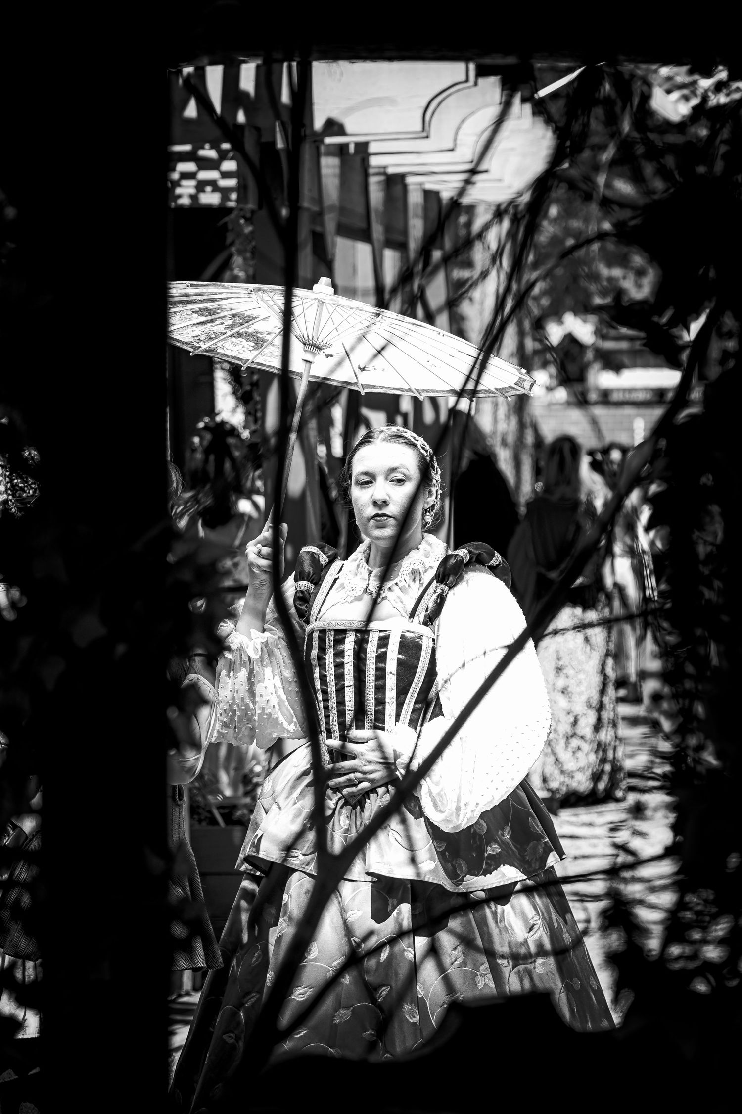Woman in colonial corset dress holding parasol framed by branches, artistic black and white portrait.