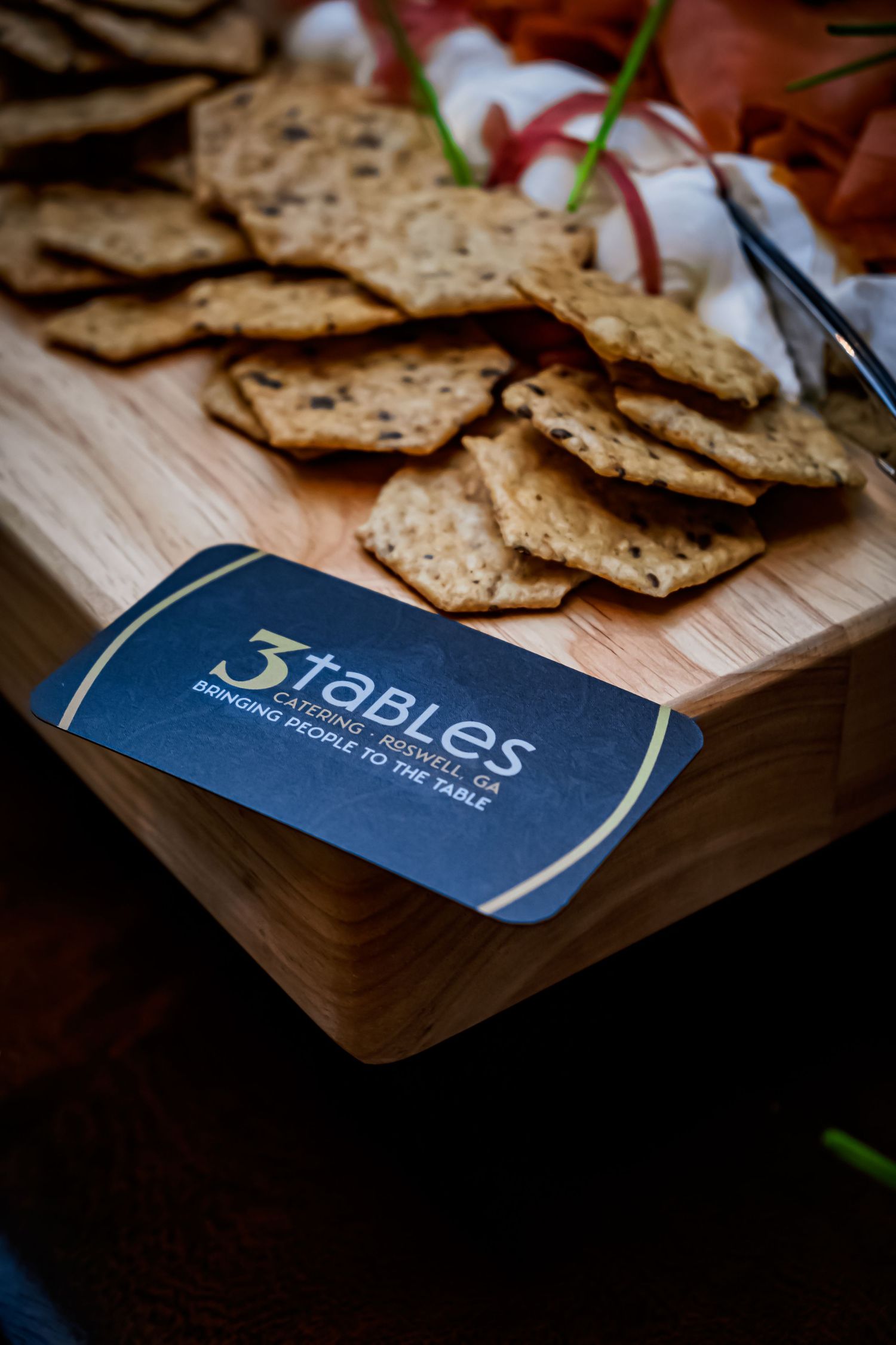 3 Tables Catering business card displayed on a wooden charcuterie board with assorted crackers and garnishes.