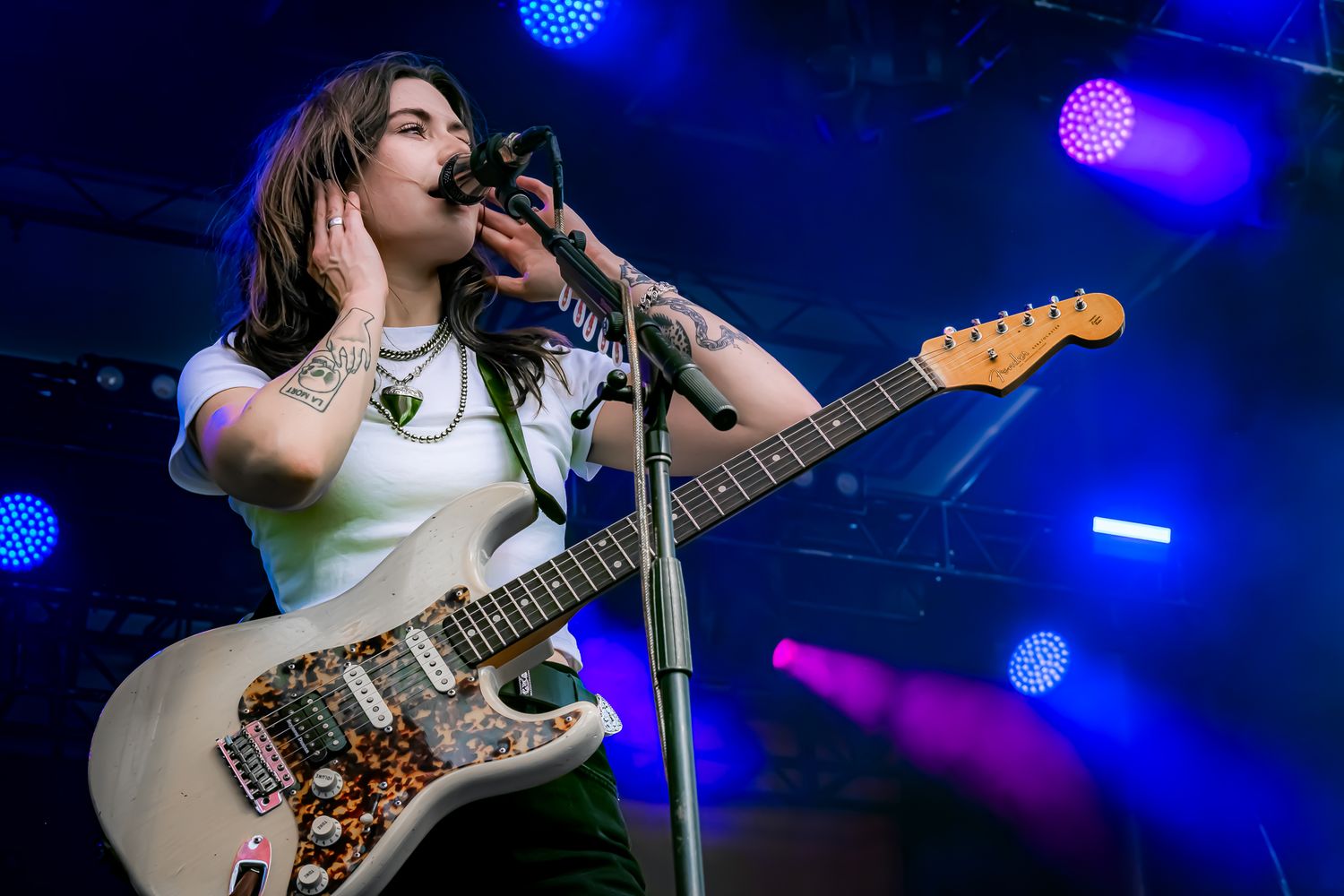 Tattooed female guitarist playing white Fender Jaguar on stage under blue and purple lights.