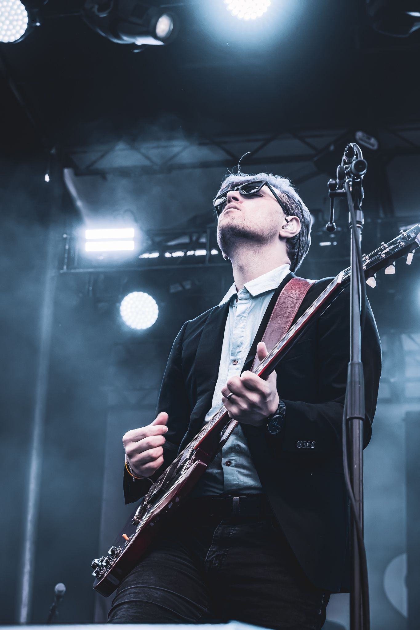 Guitarist in black suit playing red electric guitar under stage lights at live concert.