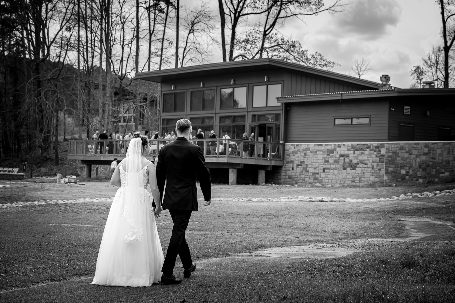 Black and white wedding photo of bride and groom walking toward a modern venue building with guests outside.