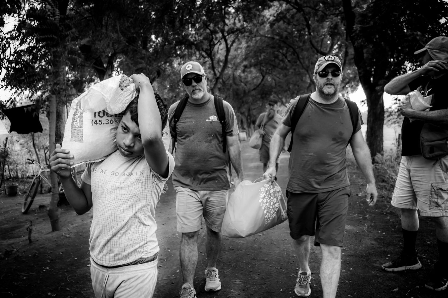 Black and white photo of men carrying heavy bags and supplies outdoors, working together in a relief or community aid effort.