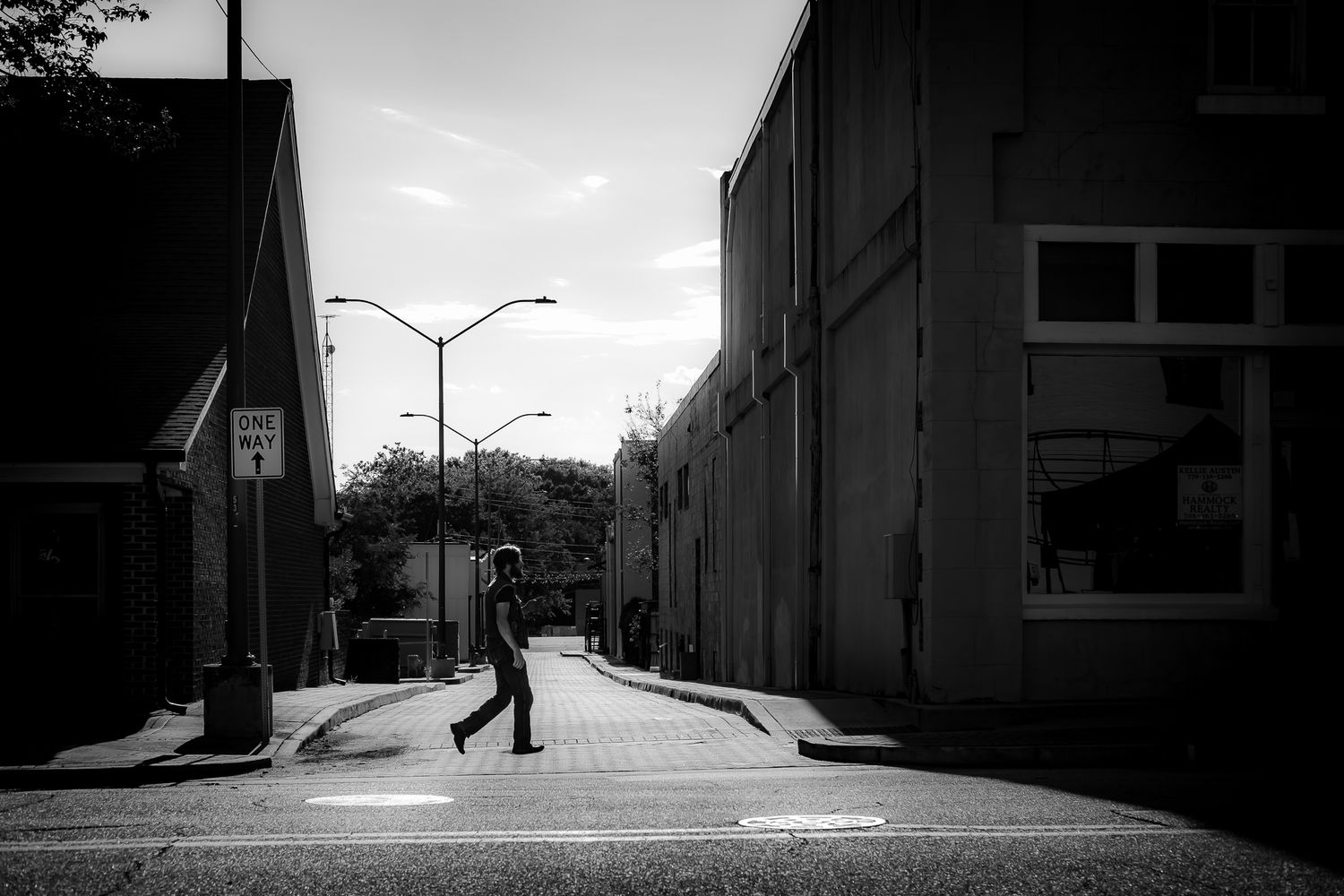 Black and white photo of a person crossing a quiet urban street with buildings and street lights in the background.
