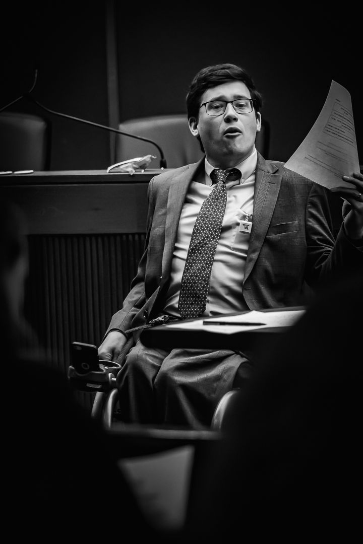 Black and white photo of a suited man with glasses holding papers while seated at a formal meeting.