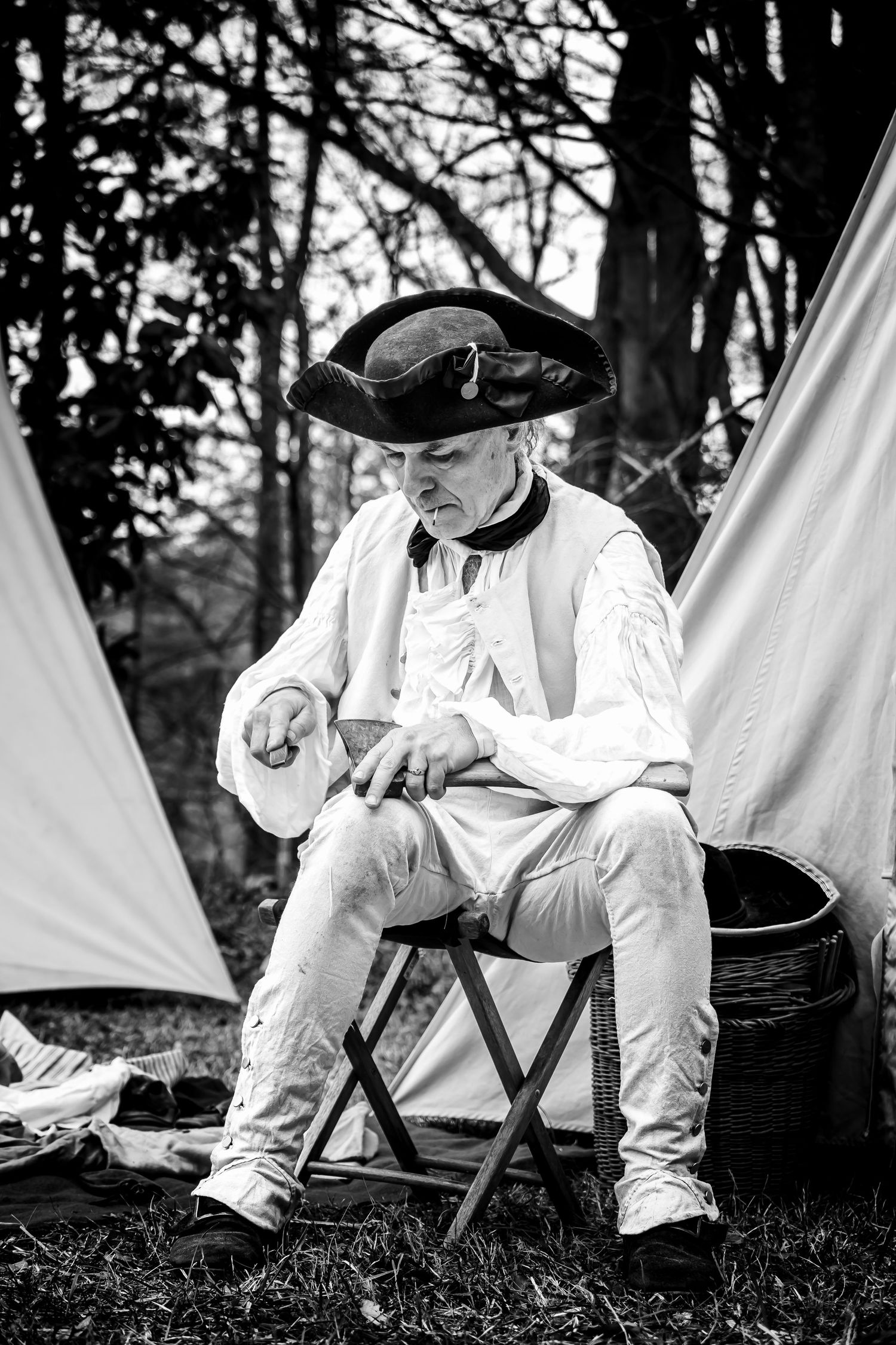 Man in tricorn hat and white colonial attire sits pensively on camp stool, black and white portrait.