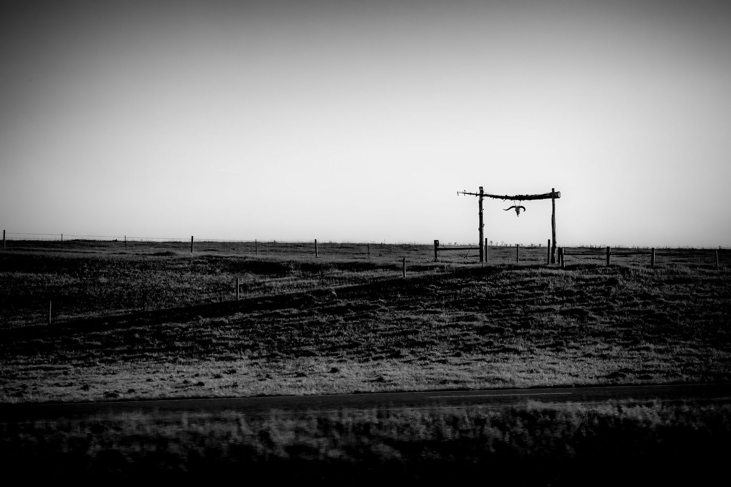 Minimalist black and white photo of a wooden ranch gate on a vast empty prairie under clear sky.